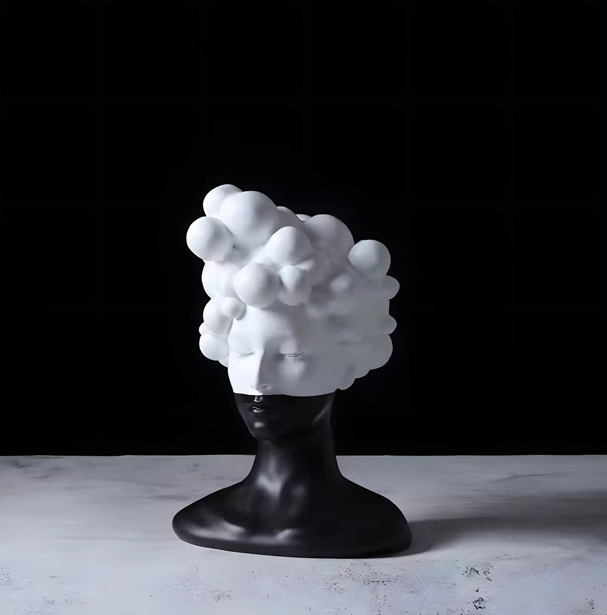 A sculptural decorative head with a glossy black neck and base supporting a white face and voluminous cloud-like hair, displayed on a light marble surface against a black background.