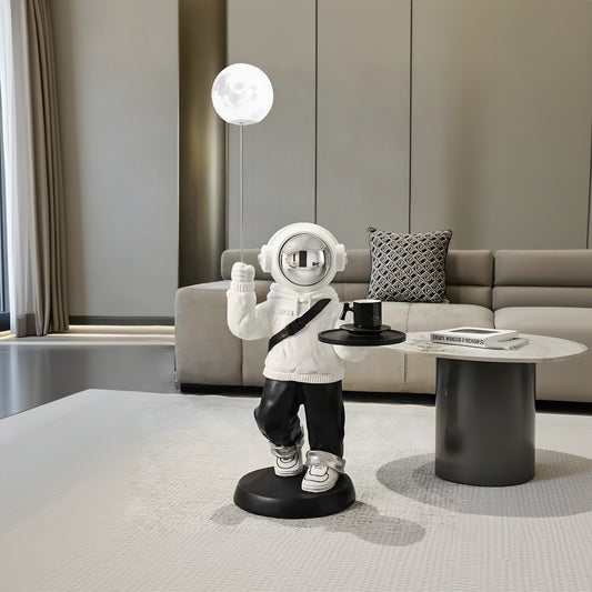 A whimsical astronaut figurine standing on a round black base, holding a small tray with a black mug and wearing a white hoodie and black pants. The astronaut is posed in a modern living room setting next to a sofa and a glowing spherical lamp.