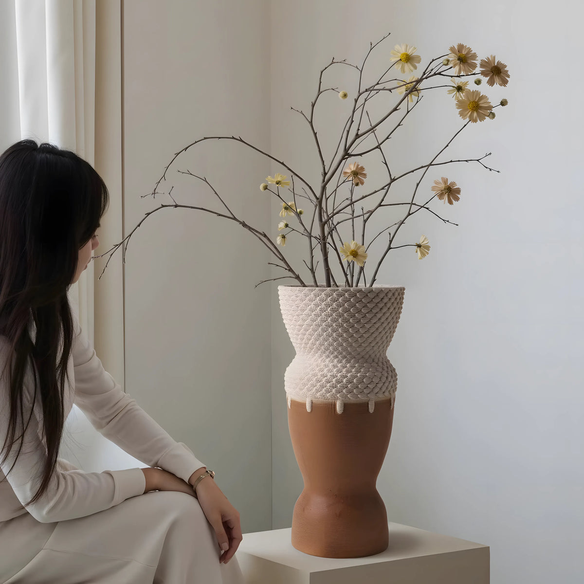 A woman in a cream outfit sits beside a sculptural ceramic vase holding dried branches with small yellow flowers, set in a softly lit neutral interior.