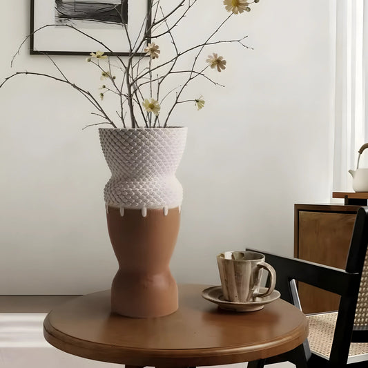A sculptural two-tone ceramic vase with a textured white top and a matte terracotta brown bottom, filled with dried branches and small flowers, displayed on a round wooden table in a styled interior setting.
