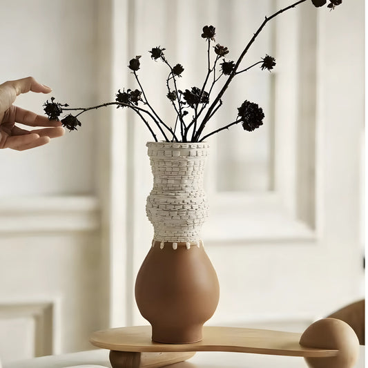 A two-tone ceramic vase with a textured white woven upper section and a matte brown rounded base, holding dried dark flower stems. A human hand is reaching in from the left, and the vase sits on a light wood table in a softly lit interior setting.