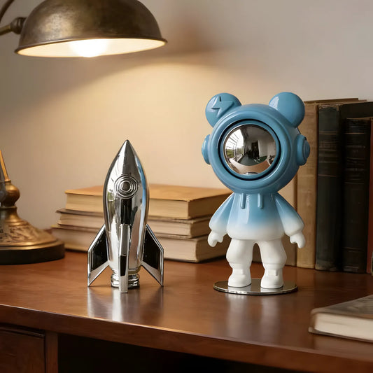 Modern glossy sky blue astronaut bear sculpture with a silver visor on a dark wooden office desk next to a silver toy rocket