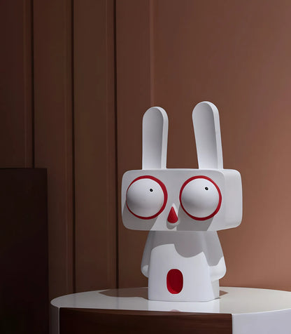 The large white pop art rabbit statue styled on a round bedside table against a warm brown minimalist paneled wall